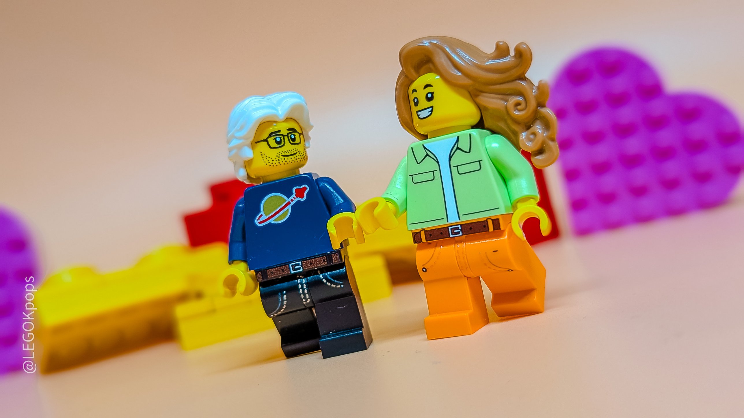 Two minifigures, one with white hair and glasses in a classic space shirt, the other with long brown hair, among colourful bricks